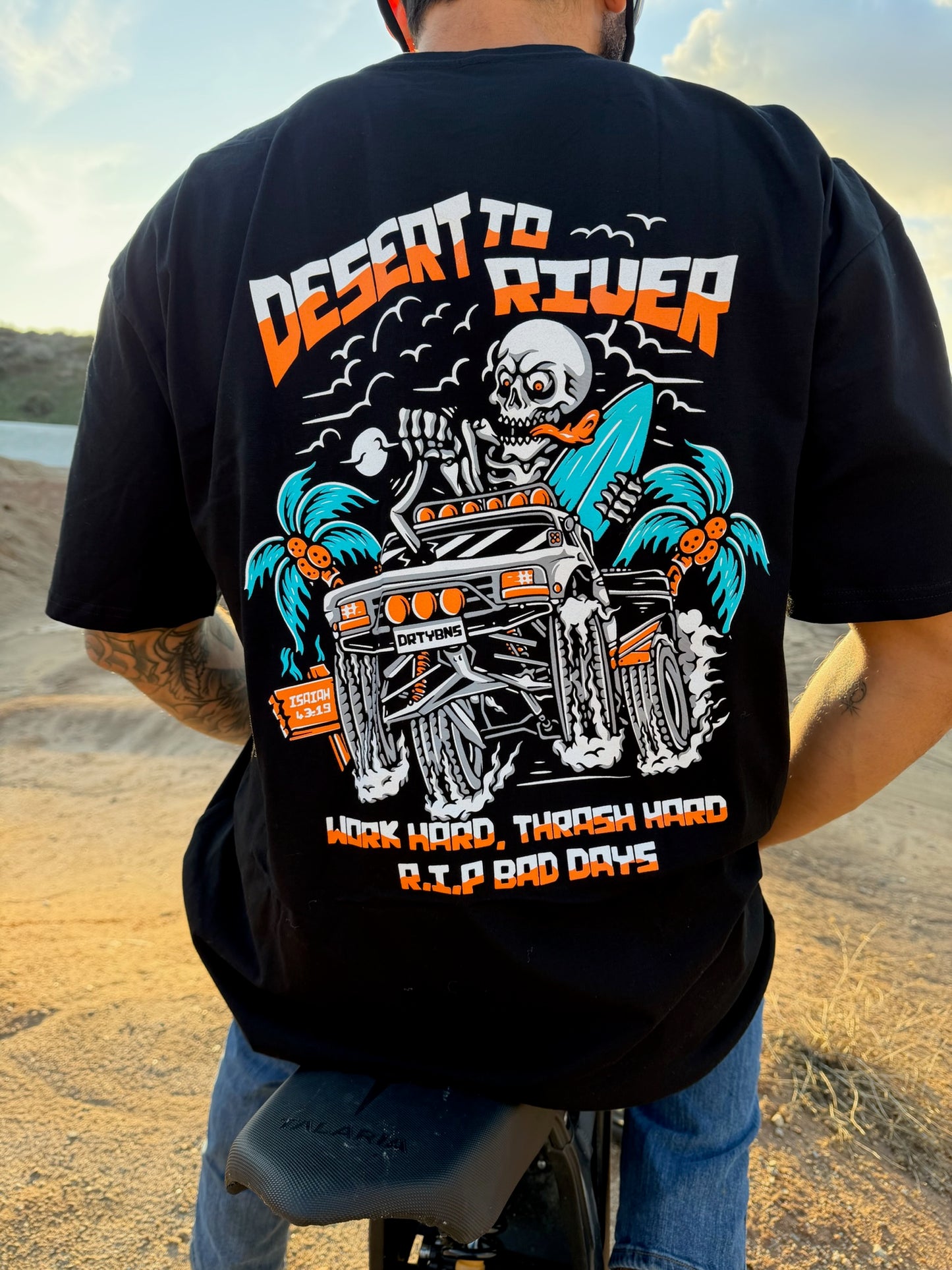 Person wearing a black t-shirt with a skeleton on a motorcycle graphic and text in a desert setting.